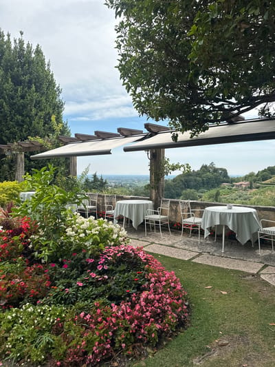 A restaurant patio overlooks a valley in Maser, Italy. White-clothed tables and chairs are set up under a pergola with white awnings. A stone wall separates the patio from the valley, which is covered in trees and a few houses. In the foreground, a variety of flowers bloom in shades of pink, white, and red. The sky is cloudy.