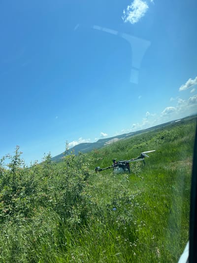 A ground-level, slightly distorted photograph, likely taken from inside a vehicle, shows a grassy field in Gorna Kremena, Bulgaria under a blue sky with scattered clouds. A piece of equipment, possibly agricultural or surveying, stands in the field, partly obscured by taller grasses and other vegetation. The land slopes gently upward in the background. The presence of a reflection in the sky confirms that the image was taken through a window.