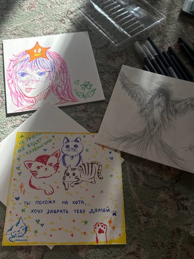 The image shows a collection of drawings and art supplies scattered on what appears to be a textured surface, possibly a rug.
There are four main paper-based artworks:
1. A drawing of wings in pencil.
2. A colorful drawing of a female character with pink hair, blue eyes, and a star on her head.
3. A postcard-style drawing with multiple cats in different colors and patterns. The background is decorated with stars, hearts, and other small motifs. There is text in Russian on the postcard.
4. A blank sheet of paper.
Art supplies are visible: a plastic container and a collection of black pens or markers.
The setting appears to be indoors. There's no indication of a specific location or connection to Uryupinsk, Russia.