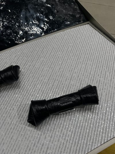 An overhead shot depicts two black, elongated objects on a textured light gray surface within a display setting in 성동구, South Korea. The prominent object, centrally positioned, is made of what appears to be black, faux-crocodilian leather, folded into a knot-like form. The word "TAMBURINS" is embossed in light lettering on its surface. A second, similar black object is partially visible in the upper-left quadrant. The light gray surface features a pattern of fine, parallel diagonal ridges and is covered with small, clear droplets or a textured, glossy clear coating, giving it a wet appearance. In the upper-left corner, a dark, highly reflective surface with an irregular, wavy texture is present, separated from the gray surface by a thin, metallic border. No individuals or active interactions are visible in the static scene.