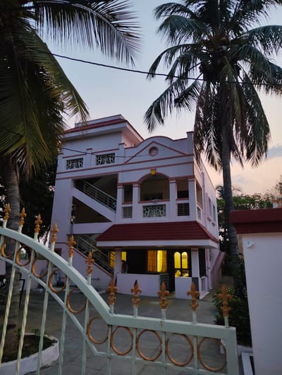 The image depicts a three-story residential building in Kammavaripalli, India, taken during twilight. The building is painted in light pink with a red tiled roof and features an external staircase on its left side. The ground floor has illuminated windows, suggesting interior lighting. A decorative white metal gate with golden accents is visible in the foreground, partially obstructing the view of the building. Two tall coconut palm trees frame the building, adding to the tropical ambiance. The sky transitions from light blue to orange, indicating the time of day. A portion of a white wall is visible on the right side of the image. No people are directly visible in the frame.