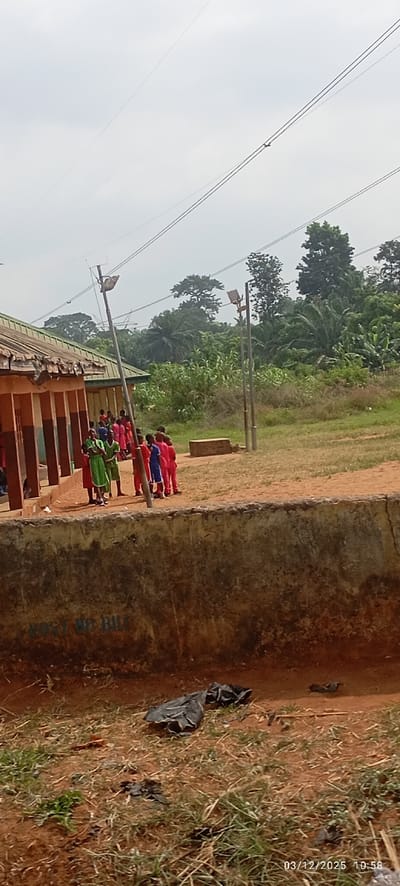 This image, captured on March 12, 2025, at 10:58, depicts an outdoor scene in Abagana, Nigeria. The foreground consists of reddish-brown dirt and sparse, dry grass, with discarded black plastic refuse visible on the ground. A weathered concrete retaining wall, stained and marked with faint blue lettering "POST NO BILL," traverses the mid-foreground.
Behind the wall, a group of approximately 10-15 individuals, likely children, stand in a line or loose formation. They are positioned next to and extending from a single-story building with a corrugated metal roof and a shadowed veranda supported by painted columns in brown and light blue/grey sections. The individuals are dressed in brightly colored clothing, primarily red/pink, with some wearing green and at least one in blue. Their exact actions are limited to standing.
Two tall, slender poles, equipped with solar panels and light fixtures, stand upright in the open dirt area to the right of the building. A rectangular concrete block rests on the ground near the base of the rightmost pole. The background features dense green vegetation, including trees and shrubs, suggesting a natural or undeveloped area. The sky is overcast or hazy, appearing light grey. Multiple thin power lines or communication cables crisscross the upper portion of the frame, with a small, dark, indistinct object suspended from one of the higher lines.