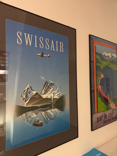 The image shows two framed posters hanging on a white wall. The poster on the left features a blue sky with a white airplane flying above snow-capped mountains reflected in a body of water. A small boat with a single person fishing is visible in the water. The word "SWISSAIR" is prominently displayed at the top of the poster in white lettering. The poster on the right has a colorful illustration of a mountainous landscape with a red bridge and a body of water. Text at the bottom of this poster reads "par le rail dans nos montag". The overall scene appears to be an interior wall, likely in a gallery or a home.
