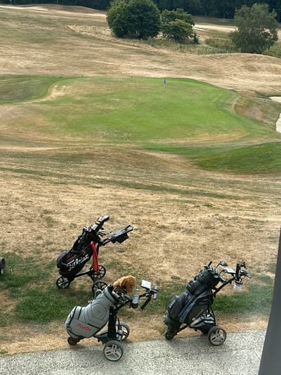 The image shows a golf course in Méan, Belgium. Three golf bags on trolleys are in the foreground. Two of the trolleys are grey and black, and one is black. One grey trolley has an "Audi" logo. The golf bags contain golf clubs. A stuffed animal is attached to one of the trolleys. The ground is dry and covered with yellowed grass, with patches of green grass on the golf course itself. Trees are visible in the background. The sky is overcast.