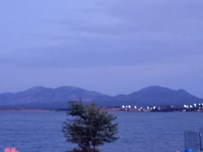 An image taken at dusk shows a body of water with a tree in the foreground. In the distance, mountains are visible, and along the shoreline, lights from a settlement twinkle. The sky is a pale, hazy blue, indicative of twilight. The scene is calm and tranquil, with no visible people or animals. The location is the Bayanaul rural district in Kazakhstan.