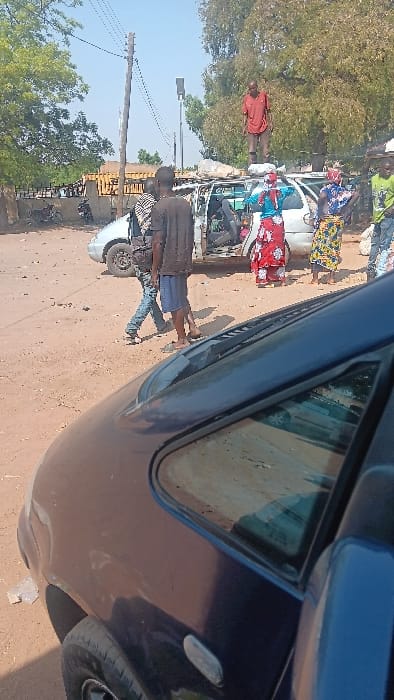 The scene is an unpaved outdoor area in Langtang, Nigeria, under a clear blue sky. A heavily damaged white minivan is centrally positioned on reddish-brown dirt ground. The minivan's roof is significantly dented, with a light-colored object resting on top. Its passenger-side doors are either missing or heavily damaged, exposing a stripped interior.
Seven individuals are visible. An adult male in a red shirt and dark shorts stands barefoot on the roof of the white minivan, facing right. To the left of the minivan, two adult males are present; one in a striped shirt with a shoulder bag walks left, while another in a dark t-shirt and shorts stands near the vehicle. To the right of the minivan, three adult females in patterned attire are standing and observing.
In the foreground, the front passenger side of a dark blue vehicle is partially visible. The background features green trees, a wooden utility pole with overhead wires, a dark grey solar street light, and a low concrete wall behind which structures and a motorbike are discernible. The overall scene suggests post-incident observation or salvage activity around the damaged vehicle.