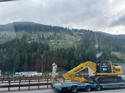 The image, taken from a vehicle, shows a Komatsu excavator loaded onto a flatbed trailer on a highway. The excavator is yellow with "KOMATSU" branding and model number "PC 170 LC" visible. The trailer is teal and black, with multiple axles and tires. In the background, a dense coniferous forest covers a steep mountainside under a cloudy sky. A power line tower and overhead wires are visible on the mountain. On the highway in the middle ground, there are several white vans, a semi-truck with "PFLUGER TRANS" branding, and other vehicles. A tall light pole with multiple lights stands on the right side of the highway. The scene appears to be in a mountainous region, possibly near Brenner - Brennero, Italy, given the presence of a highway and dense mountain forestation. There are no people clearly visible in the image.