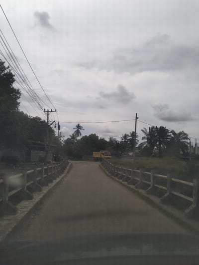 A slightly overcast, daytime, head-on view of a paved road leading towards a yellow truck. The road is flanked on both sides by a low concrete barrier with yellow railings. On the left, a dense line of dark green trees borders the barrier, with electrical poles and wires running above them. Some buildings are visible behind the trees. On the right, the barrier is bordered by a grassy area and more trees, including several palm trees. In the distance, the yellow truck is positioned on the right side of the road. The sky is filled with a mix of dark and light clouds. The overall scene depicts a semi-rural or suburban road in Pantonrayeuk Satu, Indonesia.