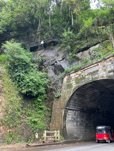 The image shows the Ramboda Tunnel in Sri Lanka. The tunnel entrance is constructed from brick and stone, and the surrounding area is covered in lush green vegetation. There is a rocky cliff face alongside the tunnel entrance. A portable generator sits on the road just inside the tunnel entrance.