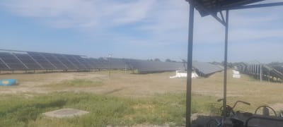 This is a wide, eye-level outdoor shot of a solar panel farm under a clear sky with scattered clouds. The solar panels are arranged in long rows, angled towards the sun. In the foreground, to the right, is a partial view of a covered structure with a dark, slatted roof and two dark support posts. A bicycle with a basket is parked to the right of the structure, and a part of a cart is visible. The ground is covered in dry, light-brown grass with patches of greener vegetation. A blue, boat-shaped object is visible in the middle ground to the left, near the solar panels, and a concrete utility cover is also present in the foreground. The background consists of a treeline and the horizon. The scene appears to be located in Ushtobe, Kazakhstan.