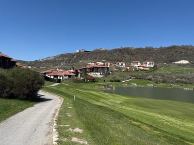 This expansive landscape photograph captures a scenic residential and recreational area in Bozhurets, Bulgaria, under a clear, bright blue sky. The setting features a verdant, hilly terrain developed with numerous buildings and a prominent water body, likely a golf course.

In the foreground, a narrow, winding asphalt path with visible cracks curves from the lower left towards the center right, bordered by green grass. A large, dense bush occupies the immediate left side, partially obscuring the view of buildings beyond. To the right of the path, manicured green fairways, characteristic of a golf course, slope down towards a calm, dark green lake or large pond in the midground. White markers are visible on the grass.

The hillsides are dotted with numerous residential buildings, primarily houses and apartment blocks, many featuring distinctive red-tiled roofs and light-colored facades. These structures are scattered across the slopes, some clustered together, others more isolated, blending into the natural environment. On the higher slopes, the vegetation becomes sparser with more dry, deciduous trees and bushes, while the areas around the golf course are lush and green. Several modern-style buildings are visible on the crests and upper slopes of the hills in the background.

The sky is a brilliant, cloudless blue, indicating sunny weather. The absence of people suggests a quiet moment, emphasizing the natural beauty and architectural integration of the resort. No visible text is present in the image. The scene portrays a serene and well-maintained resort community, likely designed for leisure and residential living, integrated into a picturesque, hilly landscape.