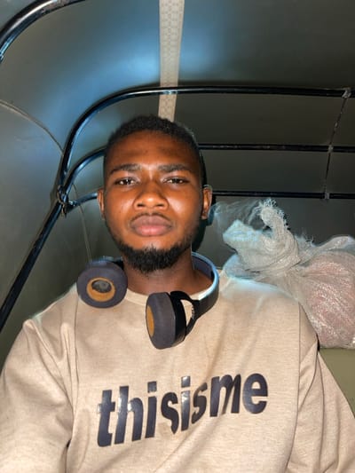 A young man with a short, dark haircut and a well-groomed beard poses for the camera. He wears a beige sweatshirt with the word "thisme" printed in large, bold black letters down the arm. Black headphones are around his neck. He is standing or sitting in what appears to be a curved metal structure with vertical bars. A white, translucent plastic bag or wrap is visible behind him near the top of the frame.