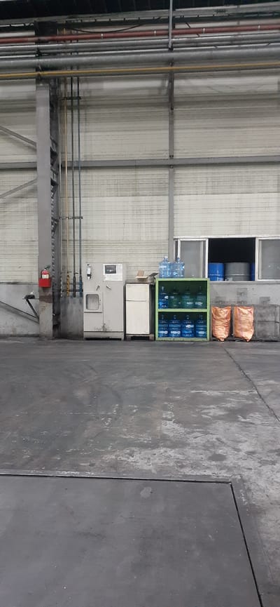 This is a rotated image of the interior of a warehouse or industrial facility in Miryang-si, South Korea. The scene is viewed from a low angle, looking upwards. On the left side of the frame, there are many vertical pipes, painted in various colors including red, yellow, and gray, running along a corrugated metal wall. These pipes are supported by a grey metal structure.
In the center of the image, there is a green shelving unit filled with blue plastic water bottles, commonly used for water dispensers. Above this unit, there are two large orange bags, possibly containing supplies or materials. Below the water bottles, there are some white cabinets and a grey utility box with a circular window. Further down, more pipes and a red emergency button are visible, attached to a metal beam.
The right side of the image shows a rough, grey concrete floor with visible seams. The overall impression is of a functional, utilitarian space. There is no indication of the time of day or weather conditions. No people are visible in the image, and no text is discernible.