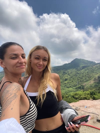 Two women are taking a selfie on a rocky outcrop overlooking a lush green mountainside with a blue sky and scattered clouds. The woman on the left has dark hair, visible tattoos on her arm and neck, and is wearing a black and white striped tank top. The woman on the right has blonde hair, is wearing a black tank top with white trim, and is holding a pink cell phone. The location is identified as Nam Hải, Vietnam.