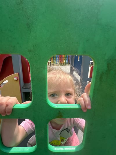 A blonde toddler with wide blue eyes peers through a green plastic structure, likely part of a playground. The child's face is partially visible, with one hand gripping the green frame and the other resting on it. The child wears a pink t-shirt with a cartoon design. In the background, a red and yellow structure, possibly a slide, is visible, along with colorful vertical elements of a playground fence. The ground appears to be paved. The scene is illuminated by daylight. The location is identified as Donchovci, Bulgaria.