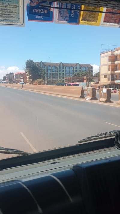 The image depicts a daytime scene from within a vehicle, looking forward through the windshield. The perspective is slightly elevated, suggesting a bus or larger vehicle. The environment is an urban setting within Kinoo ward, Kenya, as indicated by a visible flyer inside the vehicle listing "Kinoo Ward, Kiambu County" under "Nairobi Office" branches.
The foreground shows the vehicle's dark gray dashboard with a textured surface. Two windshield wipers rest horizontally on the lower portion of the windshield. Along the top edge of the windshield, several rectangular advertising flyers are affixed. These flyers display partial text such as "GITURU," "I fuel@," and "UTATAM," along with branch contact information for Nairobi, Mombasa, and Eldoret.
The midground features a multi-lane asphalt road, likely a dual carriageway, extending into the distance under a clear blue sky. A light brown concrete median barrier separates the visible lanes from oncoming traffic. Beyond this primary barrier, additional, smaller concrete barriers are irregularly spaced, some exhibiting white painted markings or graffiti. Vehicles are present on the opposing side of the road; a dark sport utility vehicle (SUV), a pink pickup truck, and a distant white sedan are visible. No individuals are distinctly identifiable on the road.
The background consists of various multi-story buildings. On the left, several buildings with reddish-brown roofs and light-colored facades line the street. Further back and centrally located, a prominent five-story building with a dark-tiled, multi-gabled roof and numerous uniformly-sized windows dominates the skyline. To the right, a multi-story building, possibly under construction or recently completed, features a lighter-colored facade with some exposed concrete elements and a flat roof partially enclosed by fencing. Sparse green trees are interspersed among the structures. The sky is uniformly clear and blue, indicating fair weather conditions.