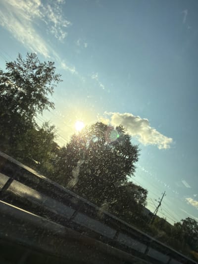 The image depicts a view through a dirty window, likely from a vehicle, looking towards a bright, sunlit sky. The sun is intensely bright, positioned slightly to the left of the center, casting a strong glare and lens flare. Silhouetted trees with dense foliage are visible against the sky, some of which are illuminated by the sunlight. Wispy white clouds are scattered across the clear blue sky. A power line pole with visible wires is present in the background, suggesting an urban or suburban environment. A dark railing or barrier is prominent in the foreground, running horizontally across the lower portion of the image. The overall scene suggests a daytime setting, possibly late afternoon due to the low angle of the sun and the warm lighting. The presence of the power pole and what appears to be a road barrier indicates a location along a transportation route. The dirt on the window obscures some details of the scene. There are no people or specific identifiable landmarks that would pinpoint a precise location within Mölndal, Sweden.