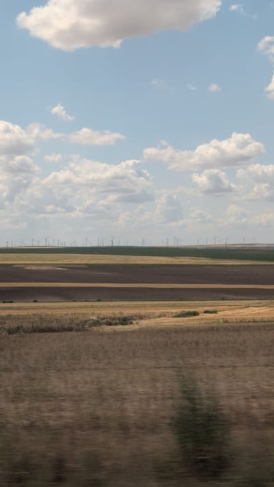 The image depicts a vast agricultural landscape under a partly cloudy sky. In the foreground, the fields are blurred due to motion, suggesting the photo was taken from a moving vehicle. The middle ground shows distinct rectangular plots of land, some appearing plowed and dark brown, others with dry, yellowish vegetation. In the distance, a large wind farm stretches across a green, rolling hill, with numerous wind turbines clearly visible. The sky is a soft blue with scattered white and gray clouds. The scene is characteristic of rural areas with renewable energy infrastructure. There is no discernible indication of the city of Baia, Romania, within the image.