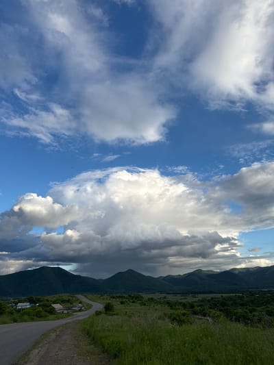 The image depicts a rural landscape under a dramatic sky with large, billowing clouds. In the foreground, a paved road curves to the left, bordered by tall green grass. To the left of the road are a few small, simple houses with blue roofs. Beyond the houses and road, rolling green hills covered in dense vegetation form the middle ground. In the distance, a series of dark green, tree-covered mountains stretch across the horizon. A rain shower is visible in the far distance, creating a visible curtain of precipitation falling from the clouds onto the mountain range. The sky is a vibrant blue, punctuated by the white and gray clouds, some of which are illuminated by sunlight, creating a contrast between light and shadow. The overall scene suggests a transitional weather event in a remote, natural setting. The location is identified as Teployozersk, Russia, which is consistent with the depicted mountainous and rural terrain. There are no people or animals visible in the image.