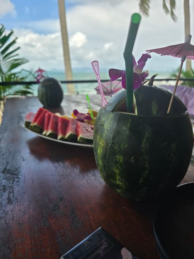 The image shows a watermelon drink served in a hollowed-out watermelon, decorated with an orchid, a paper umbrella, and a green straw. Slices of watermelon are arranged next to the watermelon drink. The drink and watermelon slices are set on a wooden table. In the background, the sea and sky are visible, along with palm trees. This image was likely taken in Ban Khlong Hia, Thailand, given the location tag. A small item with printed text is placed on the right of the watermelon.