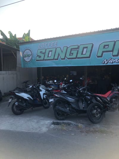 The image depicts the exterior of a coffee shop named "SONGO PI" in Kalidawir, Indonesia. A large teal banner with the text "COFFEE SONGO PI" in green lettering hangs above the entrance. Below the banner, several motorcycles are parked on a concrete surface. Some of the motorcycles are white and black, while others are black and red. In the background, the interior of the shop is visible, with people gathered inside. Banana trees are also visible to the left of the shop. The lighting suggests it is daytime, possibly late afternoon, with the sun casting a warm glow.