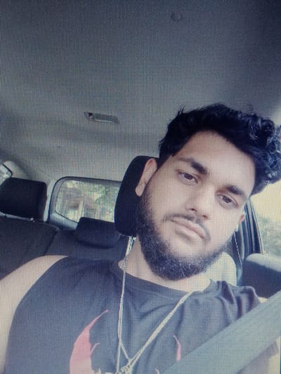 The image shows a medium shot of a young man with dark, curly hair and a beard, sitting in the driver's seat of a car. He is wearing a black sleeveless shirt and a silver chain. The man is looking directly at the camera with a neutral expression. The interior of the car is visible, including the headrests of the front seats and the back seats, as well as the side window. Outside the car, trees and foliage are visible through the window, suggesting a natural or suburban setting. The image appears to be taken during the daytime. The location in Gorno Aleksandrovo, Bulgaria is not discernible from the visual information provided in the image.