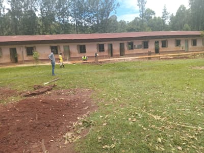 The image depicts a single-story building with a corrugated metal roof, located in what appears to be Idakho South ward, Kenya. The building has multiple windows and doors, with a weathered exterior. In the foreground, there are three individuals: one standing, one bending over, and one wearing a yellow shirt. There are piles of dirt and felled trees present on the grassy terrain. A yellow rope is visible across the grass. In the background, there are trees. The environment suggests a rural or semi-rural setting. Construction or renovation activity is indicated by the presence of lumber and work being done on the building.
