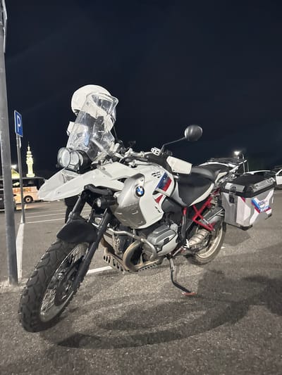 An outdoor, eye-level, medium shot captures a white BMW R1200GS Rallye motorcycle parked on asphalt in a parking lot at night. The motorcycle is positioned facing the viewer's left, angled slightly towards the camera. Its front wheel is on the left, and the rear wheel is on the right. A white helmet with a clear visor is perched on top of the motorcycle's windshield. The motorcycle is equipped with saddlebags on both sides. In the background, to the left, a tall, slender minaret of a mosque and a white bus with lettering are visible. A blue and white parking sign is mounted on a pole to the left of the motorcycle. The scene is illuminated by artificial light, casting shadows on the ground. The sky above is dark and clear. The general location is the city of Privolzhsky, Russia.