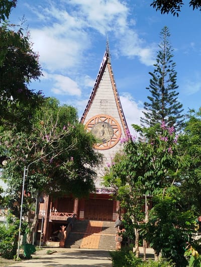 The image depicts a building with a unique architectural design in An Phu Ward, Vietnam. The building has a tall, triangular facade with a central circular motif featuring a religious figure. A series of steps lead up to the building's entrance. Trees and foliage partially obscure the view of the structure. The sky above is blue with scattered clouds. A green trash can is visible on the left side of the image.
