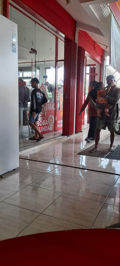 The image shows the exterior of a shop with red and white signage and red folding doors. Inside the shop, there are display cases visible through the glass. A young man in a black long-sleeved shirt and denim shorts is standing near the entrance, looking into the shop. Further inside, a woman in a black hijab and orange shirt is holding a baby, and a man in a gray hoodie and green shorts is standing next to them. The floor is tiled and reflects the interior of the shop and the people. The scene appears to be in Crewek, Indonesia, likely a shop selling food or beverages, given the "BOBA SUNDAE" signage.