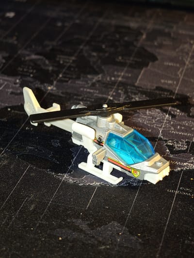 A toy helicopter, primarily silver and white with a blue canopy, is positioned on a dark surface printed with a world map. The helicopter features rotating blades, landing skids, and decals that include "75" and "METRO RESCUE" with a red "R" logo. The background map shows country outlines and text, including partially visible place names such as "NORWAY", "SWEDEN", "POLAND", and "GERMANY", with faint lines crisscrossing the surface. The lighting casts shadows from the helicopter onto the map. No people or actions are depicted. The scene appears to be a still life arrangement.
