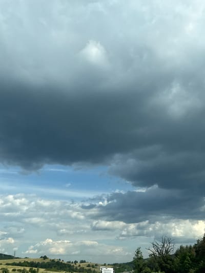 The image depicts a cloudy sky over a rural landscape, possibly in or near Rajana, Bulgaria. The upper portion of the frame is dominated by dark, ominous storm clouds, while the lower section shows a brighter sky with scattered cumulus clouds. The terrain below is hilly and covered with greenery, including trees and some dry grass. A power line tower is visible on the left side of the image, and a white truck with some text on it is partially visible at the bottom. The scene appears to be captured from a moving vehicle, suggested by the slight blur and the perspective.