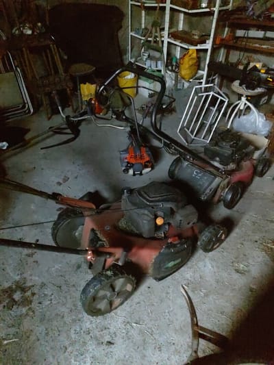 The image shows a collection of outdoor equipment, likely stored in a garage or shed. The primary subjects are two lawnmowers and a tiller. One lawnmower is predominantly red and black, with its wheels clearly visible. It appears to be a walk-behind model, tipped on its side. The other lawnmower is mostly obscured by shadows but seems to be a similar type. The tiller is orange and black, also on its side, with its tines visible.
Other objects in the scene include a yellow bucket, a white metal rack or cage, and various pieces of metal tubing and frames. There are also some plastic bags and what appears to be a yellow tarp or bag. The ground is a concrete or dirt floor, with some scattered debris and straw.
The lighting suggests an indoor setting, possibly dimly lit. There are no people visible in the image. The overall impression is one of storage or disarray, with the equipment not currently in use. The presence of lawnmowers and a tiller indicates a location where yard work is performed. The context provided, Kochmar, Bulgaria, suggests this could be a rural or suburban setting.
Visible text on the red lawnmower includes the number "75" on the engine housing. There is also some unreadable text on the yellow bag. The time of day and weather are not discernible from the image.