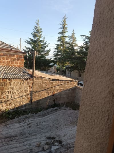 The image depicts an outdoor scene in the city of Talin, Armenia, under a clear blue sky. In the foreground, a textured, light brown wall partially obscures the view. Beyond this wall, a corrugated metal roof covers a section of a building constructed from rough-hewn, reddish-brown bricks. Adjacent to this is another building with a flat, light-colored roof, also constructed from rough stone blocks.
Several tall evergreen trees with lush green foliage are visible in the middle ground, some reaching towards the sky. In the background, a light-colored house with windows can be seen, and a white car is parked in front of it. Utility poles and electrical wires crisscross the scene, indicating a residential or semi-urban environment. The ground in the lower part of the image appears to be covered with rough sand or dirt, with some scattered stones. The lighting suggests it is daytime, possibly late afternoon or early morning, given the long shadows cast on the brick wall. There are no people visible in the image.