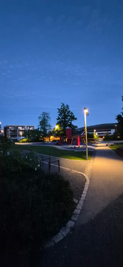 This image depicts a park area at dusk in Lillehammer, Norway. The sky is a deep blue with wispy clouds. In the background, modern apartment buildings with illuminated windows are visible. A playground with red structures and a slide stands in the middle ground, surrounded by trees. A streetlamp illuminates a pathway that curves through the park, casting a warm glow. The foreground features dark green bushes and a low metal bar structure, possibly part of an outdoor gym. The overall scene is tranquil and quiet, suggesting the end of a day.