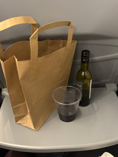 The scene displays an arrangement of objects on a light grey folding tray table, typical of a transport setting, located in Stoke-on-Trent, United Kingdom. A brown paper bag with two integrated handles occupies the left foreground, standing upright. To its right, a small, dark green glass bottle with a black screw cap is positioned, its contents partially depleted. Directly beside the bottle, a clear plastic cup holds a quantity of dark red liquid. The background is a uniformly plain, light grey interior panel. No individuals are depicted.