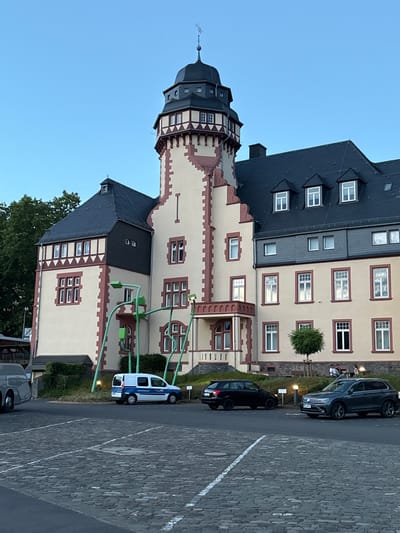The image shows a building with a distinctive tower and a red and cream facade, located in Bernkastel-Kues, Germany. The building appears to be an older, possibly historical structure. In front of the building, there is a parking lot paved with cobblestones. Several cars are parked in the lot, including a white van with "ORDNUNGSAMT" written on its side. There are also a few people sitting on benches outside the building. The sky is clear and blue, suggesting it is daytime. The overall scene depicts a public space with a prominent architectural landmark.
