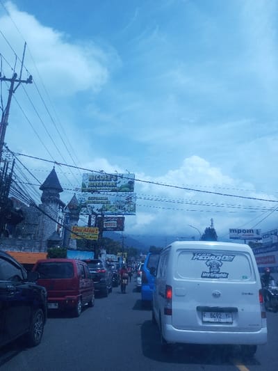 The image portrays a congested urban street scene, depicted as being in the city of Chori, Nigeria. The sky is blue with scattered white and gray clouds, indicating daytime conditions.
In the left foreground, a dark vehicle is partially visible, showing its front wheel and side. To its right, a red compact van (license plate partially visible, ending in "BL") is in a line of traffic. The paved road appears to be a multi-lane thoroughfare with a faded yellow line marking the right edge.
In the midground on the left, a multi-story building constructed with gray stone-like material features prominent turret-like structures with pointed roofs, giving it the appearance of a medieval castle or themed attraction. Above this building, a large horizontal billboard displays "NICOLE'S RIVER PARK" with cartoon graphics, accompanied by text in an Indonesian language. Below this, another sign reads "Sungei Alam 2," also featuring Indonesian text. A dense tangle of overhead power lines and utility poles runs across the upper midground.
Further down the street, a queue of vehicles including a blue van, black cars, and a red motorcycle is visible. A solitary pedestrian walks along the right side of the road, carrying items. On the far right, the rear of a white Daihatsu Gran Max van is prominently visible. It features "TEAM PEDRO 02" and "Jay GraphiX" decals on its rear window, and its license plate reads "F 8492 YF" with a validity date of "01-24". Additional commercial signage, including one with "mpoint," is visible along the right side of the street in the distance. Hazy mountains are discernible in the background beneath the cloudy sky, suggesting a mountainous or hilly environment.