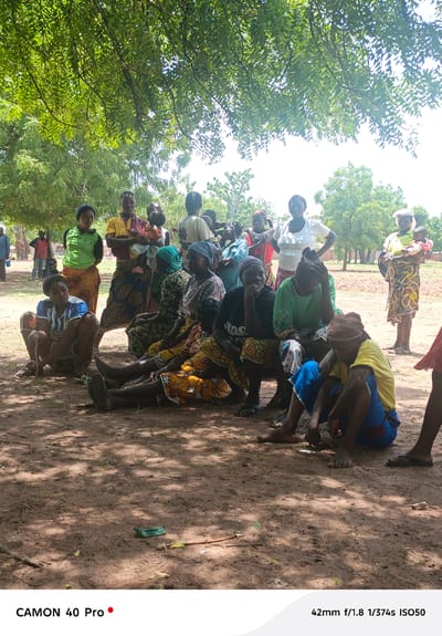 The scene depicts a group of approximately 15 women and several children gathered in an outdoor setting, likely in Gidan Kogi, Nigeria. The group is situated in a dirt clearing under the shade of a large tree. Most of the women are seated on the ground, while a few stand. Some women are holding babies or young children. Clothing appears to be a mix of Western-style shirts and traditional African patterned skirts and wraps. The background reveals scattered trees and open land, suggesting a rural location. There are a few men standing in the background. A small green object is lying on the ground in the foreground.