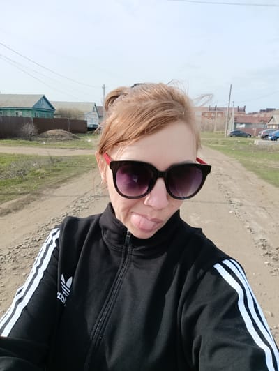 A middle-aged woman with auburn hair in a bun is standing on a dirt road in Новоалександровка, Russia. She is wearing a black Adidas tracksuit jacket with white stripes down the sleeves, and large dark sunglasses with red frames. Her tongue is sticking out slightly.
Behind her are various buildings, including a blue and green wooden house with a gray roof, and other houses with brown roofs. Several parked cars are visible further down the road. The sky is overcast.
