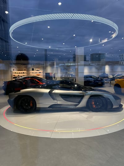 The image depicts a McLaren car showroom, likely the McLaren Glattbrugg dealership in Glattbrugg, Switzerland. Multiple McLaren cars are visible through a glass facade. The primary subject is a grey McLaren sports car positioned on a circular platform with integrated lighting. Behind it and to the left is a reflection of an illuminated ring fixture from the showroom's interior. Further back and towards the middle are two more cars, one black and the other a yellow-orange color. Interior displays with various items are visible in the background behind the cars. Exterior buildings and structures are reflected on the glass storefront, indicating an urban or suburban setting. No people are directly visible in the foreground.