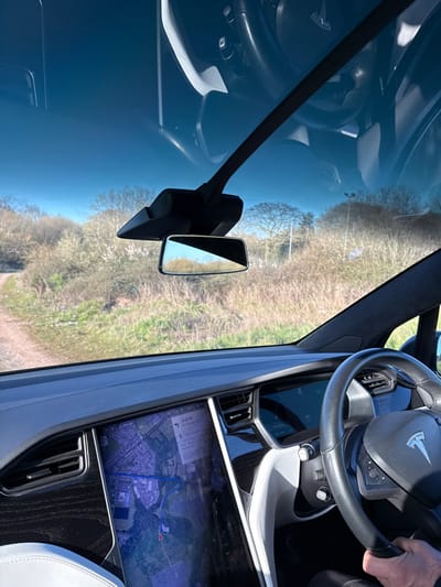 The image depicts the interior perspective from a Tesla Model X, looking forward through its panoramic windshield. The dashboard is dark grey with a wood-grain trim section on the passenger side. A large central touchscreen displays a satellite navigation map with a blue route line, indicating "0.2 mi" to the next turn and explicitly showing "Portswood" and "Swaythling Road, Southampton," United Kingdom. A human hand grips the black leather steering wheel on the right. Outside the vehicle, a dirt track is visible to the left, bordered by dry grass, sparse vegetation, and bare deciduous trees under a clear blue sky, suggesting a non-urban, possibly parkland or undeveloped area within Southampton. A thin pole is discernible in the distant foliage to the right.