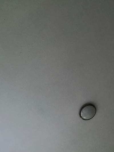 The image presents an indoor scene, likely a ceiling or wall, located in Thüngersheim, Germany. The main subject is a single, circular, surface-mounted light fixture positioned in the upper right quadrant of the frame. This fixture features a translucent or opaque white, dome-shaped cover designed to diffuse light. A distinct dark, possibly metallic, rim encircles the edge of the light's cover, attaching it securely to the surface.
The surrounding surface is uniform in color, appearing as a muted grey or off-white, and exhibits a fine, consistent texture, suggesting a painted plaster or similar finish. There are no other objects, people, or visible activities taking place in the image. The lighting is even, typical of an interior space, but does not provide cues regarding the time of day or external weather. No text is visible anywhere in the media file. The cropped hints confirm the subtle texture of the wall/ceiling and offer closer detail of the light fixture's design and its mounting ring. The scene is static, offering a close-up view of an architectural detail within a building.