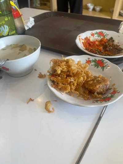 In a restaurant in Cijantra, Indonesia, a close-up, eye-level shot captures a table setting with partially eaten food. The focus is on two small white plates with a floral red pattern. One plate holds a pile of crispy, golden-brown fried food, possibly tempura or fried vegetables, with a single, round patty of what appears to be meat on the side. The second plate contains a red chili-based sauce with visible pieces of green vegetables and possibly some ground meat, indicating a spicy dish.

To the left, a white bowl contains a light-colored soup with chunks of what look like potatoes or taro, and a spoon rests in it. A green glass bottle with yellow text, likely a beverage, is also visible on the left, next to a small red-capped bottle that might contain soy sauce or another condiment. A dark brown plastic tray is partially visible in the background, suggesting the meal was served on it. The table surface is white and appears to be laminate or similar material. Several small, crispy pieces of fried food are scattered on the table around the plates. The lighting is bright, casting subtle shadows. The overall impression is that of a casual meal in progress.