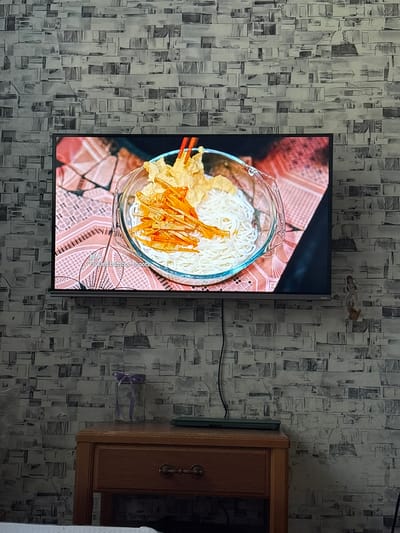 The image displays a flat-screen television mounted on a patterned wallpapered wall. The television screen shows a close-up of a bowl of noodles with some fried toppings and chopsticks. The bowl is placed on a pink, patterned surface. To the right of the television, a wooden cabinet with a dark interior is visible. A purple ribbon is tied around a rectangular object on the wall between the television and the cabinet. The lighting suggests an indoor setting, likely during the daytime given the overall brightness of the scene, though no specific time of day can be determined. The wallpaper pattern is abstract, featuring a mosaic of grey and white rectangular shapes. No people are visible in the image, and no specific weather conditions can be inferred. The location is identified as Tarasovsky, Russia. There is visible text on the television screen: "Pickled bamboo shoots".