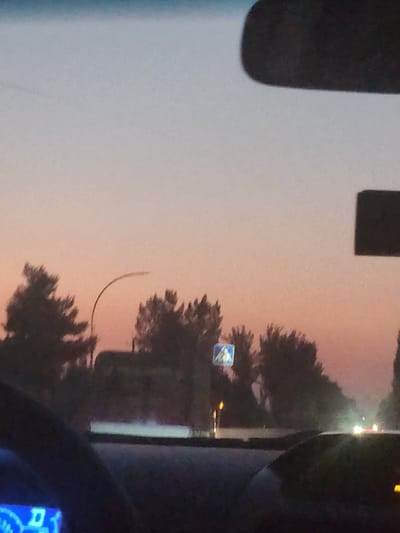 The image depicts a street scene at dusk, likely in Sariqmirzachek, Uzbekistan. The sky exhibits a gradient of soft pink and pale blue hues, characteristic of twilight. In the foreground, the dashboard of a vehicle is visible, with a blue digital display showing some numbers and symbols. A rearview mirror, dark and rectangular, is positioned at the top of the frame, reflecting the sky.
The middle ground features a row of silhouetted trees, their shapes softened by the low light. A street light with a curved pole is visible among the trees. A pedestrian crossing sign, a blue square with a white pictogram of a walking person, is also present on the left side of the image. Beyond the trees, the road is dimly lit, with the headlights of approaching vehicles creating streaks of light, suggesting traffic on a street or highway. The overall impression is one of a quiet evening on the outskirts of a city.