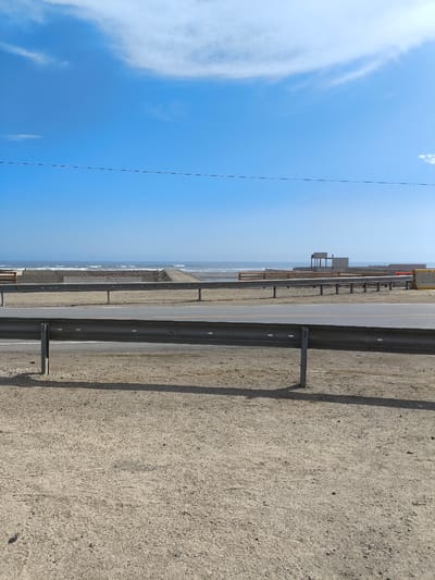 This image, likely taken in Huanchaco, Peru, shows a coastal scene. The foreground features a sandy or gravelly ground. A metal guardrail runs parallel to a road, which lies between the viewer and the beach. Beyond the road, a stretch of beach leads to the ocean, where waves are visible. A concrete structure is present on the beach, potentially a building under construction or a coastal defense. The sky is blue with some scattered clouds. A utility line is visible running horizontally across the sky.