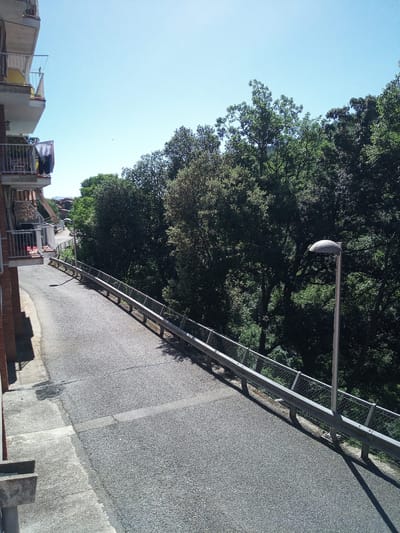The image depicts a paved road winding through a dense green forest under a clear blue sky. On the left side of the road, a building with balconies is visible, with laundry hanging on one of the balconies. A metal guardrail runs along the right side of the road, separating it from the forested area. A modern street lamp with a dome-shaped fixture stands beside the guardrail. The overall scene suggests a residential area situated alongside a natural landscape. The question implies a connection to Castellfollit de la Roca, Spain, a town known for its dramatic cliffside location, but the image itself does not contain any direct visual cues to confirm this specific location.