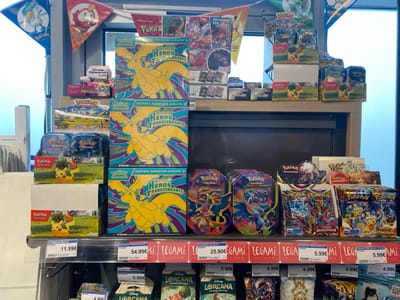 The image depicts a retail display of trading card game products inside a store in Vern-sur-Seiche, France. No individuals are visible.
The scene focuses on a multi-tiered shelving unit against a wall, with a bright window visible in the background on the right. The top tier features a stack of three large "COFFRET DRESSEUR D'ÉLITE HEROES TRANSCENDANTS" (Elite Trainer Box Transcendent Heroes) Pokémon boxes, each displaying a yellow dragon-like Pokémon against a blue and purple geometric background. These boxes are priced at 54.99€ each. To the left of these, two smaller Pokémon tins with Pikachu imagery are stacked, priced at 11.99€. Additional assorted Pokémon booster boxes and tins are arranged on the upper shelves. Triangular pennants featuring various Pokémon characters, including Quaxly and Fuecoco, are strung above the display.
The lower, prominent shelf presents a variety of Pokémon trading card products. On the far left, two distinctively patterned metal Pokémon tins are displayed, priced at 25.90€ each. Adjacent to these are several booster displays for "Pokémon Épée et Bouclier Origine Perdue" (Pokémon Sword & Shield Lost Origin) and other sets. To the right, multiple booster bundles from "Écarlate et Violet Forces Intrépides" (Scarlet & Violet Paradox Rift) and "Écarlate et Violet Flammes Obsidiennes" (Scarlet & Violet Obsidian Flames) are arranged, individually priced at 5.99€. Below this main shelf, another shelf contains smaller packaged products, including several "Disney LORCANA" starter packs and booster packs, priced at 5.90€ and 4.99€ respectively. The shelf dividers bear the "LEGAMI MILANO" brand. All prices are displayed in Euros.