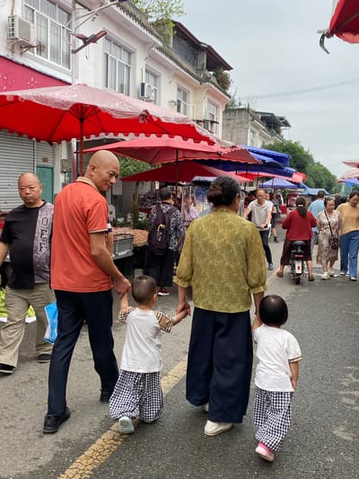 The image depicts a street scene in Chongzhou City, China, likely a marketplace. In the foreground, a man in an orange polo shirt is holding the hand of a young child wearing a plaid outfit. Beside them, a second child in a similar outfit is also being held by the man. To the left, another man in a black t-shirt stands, looking forward. In the background, various people are walking, and market stalls with red and blue umbrellas are visible. A woman in a green shirt and dark pants walks away from the camera, holding the hand of the second child. Further in the background, a woman rides a scooter. Buildings line the street, and the sky is overcast.